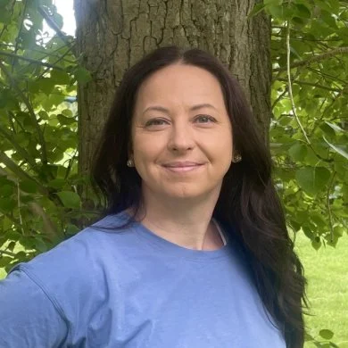 Rhonda Ortman in a blue top, standing in front of a tree