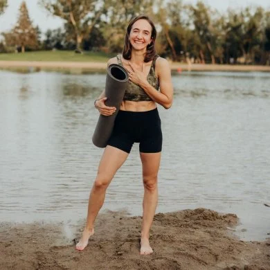 Viktoriia Nekhaieva standing barefoot on a lakeshore, holding a rolled yoga mat